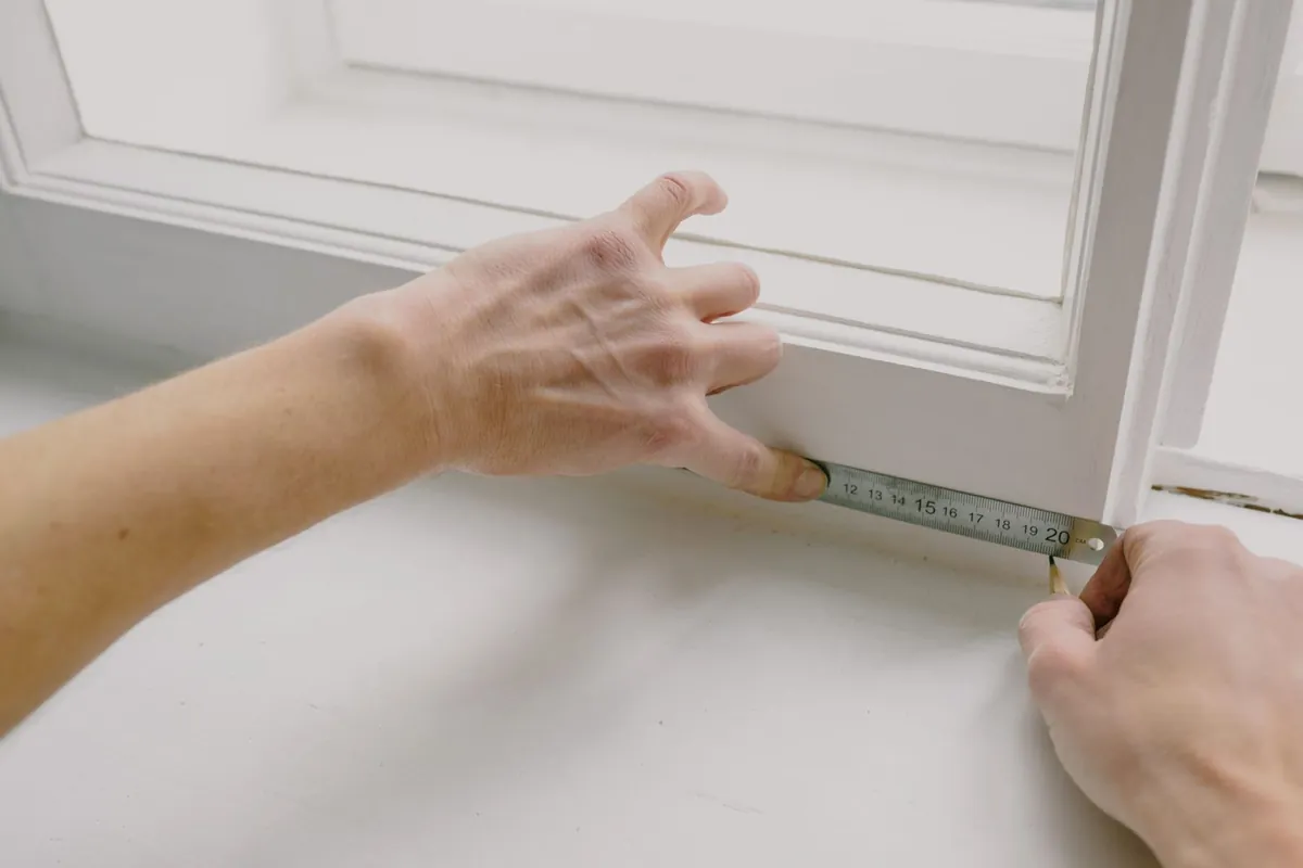 Hands measuring a white window frame with a tape measure